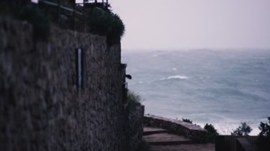 Storm waves hit the rocks in a small bay with a small coastal village. Stone path along the sea. Dramatic sea. Stormy weather. White sea foam on the stones. Mediterranean stone village.