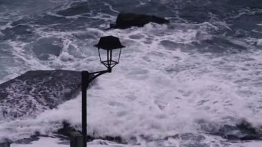 Storm waves hit the rocks at the stone embankment. Vintage lantern in the foreground. Dramatic sea. Storm. White sea foam on the stones.