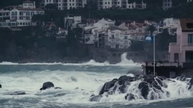 Storm waves hit the rocks in a small bay with a small coastal village. Dramatic sea. Stormy weather. White sea foam on the stones. Mediterranean white village houses on the background.