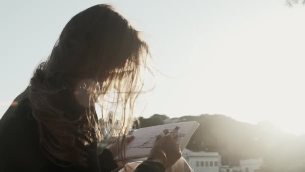 A művész rajzokat készít a napos sétányon. A lány tengerparti tájat rajzol fehér mediterrán házakkal. Fekete-fehér rajz béléssel fehér papíron. Arany napfény. Rajzolás a szabadban. — Stock videók
