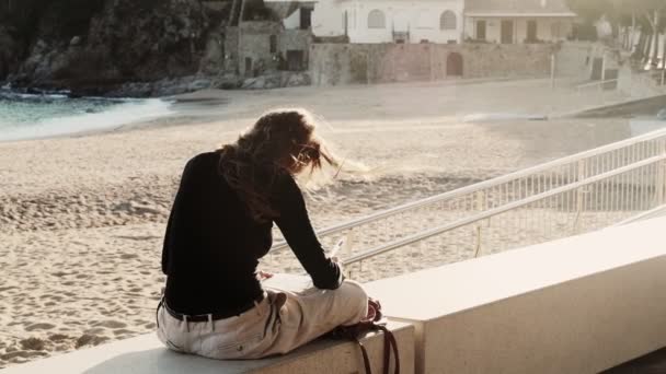 Artysta robi szkice na słonecznej promenadzie. Dziewczyna rysuje nadmorski krajobraz z białymi śródziemnomorskimi domami. Czarno-biały rysunek na białym papierze. Złote słońce. Rysunek na zewnątrz. — Wideo stockowe