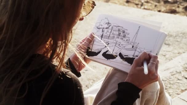 The artist makes sketches on the sunny promenade. Girl draws a coastal landscape with fishing boats. Black and white drawing by liner on white paper. Golden sunlight. Drawing outdoors. Hand drawing. — ストック動画