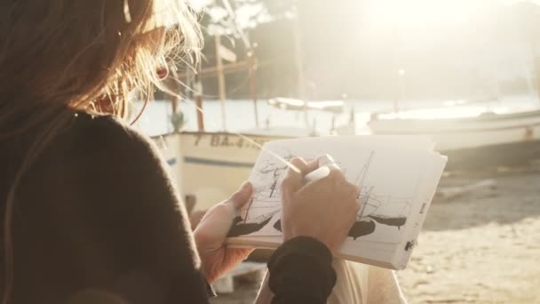 De kunstenaar maakt schetsen op de zonnige promenade. Meisje tekent een kustlandschap met witte mediterrane huizen. Zwart-wit tekening door voering op wit papier. Gouden zonlicht. Buiten tekenen. — Stockvideo