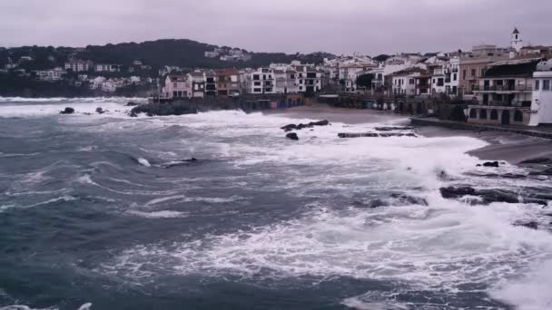 Storm waves hit the rocks in a small bay with a small coastal village. Dramatic sea. Stormy weather. White sea foam on the stones. Mediterranean white village houses on the background. — Stok video
