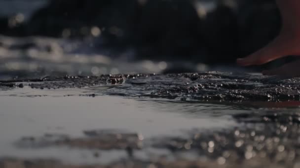 Female bare feet go along the coastal cliffs and step into puddles of water. A splash of water underfoot. Vacations and summer carefree. Walk along the beach. Waves and sea foam in the background. — Stok video