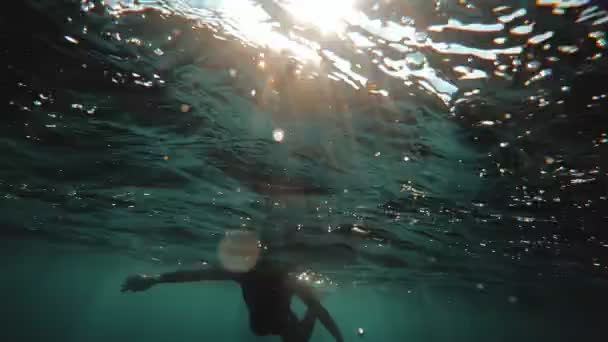 Redhead girl in a black neoprene suit splashes and swims underwater. Sun rays in clear emerald water. Spray and sun glare on the sea sand bottom. Swimming in open water. Beautiful underwater landscape — Stockvideo