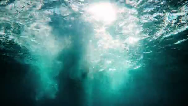 Redhead girl in a black neoprene suit splashes and swims underwater. Sun rays in clear emerald water. Spray and sun glare on the sea sand bottom. Swimming in open water. Beautiful underwater landscape — Stok video