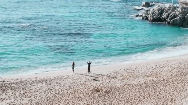 A couple in a black neoprene suits ready to swim and enters the azure water. Sun rays in clear emerald water in beautiful bay. Spray and sun glare. Swimming in open water. Triathlon training. — Stock Video