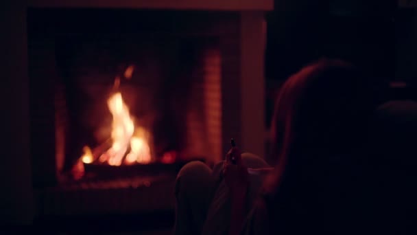 Rear view of a girl sitting in front of a fireplace with the smartphone. A wonderful evening by the burning fireplace. Warmth and comfort. Warm glare from the fire. Relaxing at home. Winter evening. — 비디오