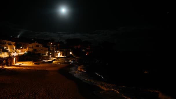 Vista noturna de uma pequena vila piscatória mediterrânea. Reflexão da lua tola na água. Luzes noturnas à beira-mar. Ondas salpicos em uma pequena baía com uma pequena aldeia costeira. Noite mar branco casas — Vídeo de Stock