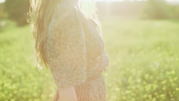 Dziewczyna w zielonej sukience stojąca na polu głupcem kwitnących żółtych kwiatów. Długie włosy kobieta podziwia widok na piękną wieś. Złote światło w sielankowym krajobrazie. Spokój i spokój. — Wideo stockowe