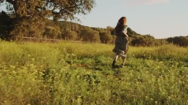 Chica en vestido verde camina corriendo por los campos tonta de flores amarillas florecientes. Mujer de pelo largo corriendo por el hermoso campo. Luz dorada en un paisaje idílico. Paz y tranquilidad — Vídeo de stock