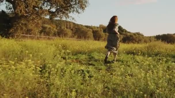 Girl in green dress walks running through the fields fool of blooming yellow flowers. Long hair woman running around the beautiful countryside. Golden light in idyllic landscape. Peace and tranquility — ストック動画