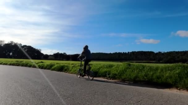 Una niña de pelo largo monta en bicicleta en un camino rural entre campos, bosques y prados. Un paisaje pintoresco. Paseo en bicicleta rural. Callejuelas, bosques con pinos y cipreses. Luz dorada . — Vídeos de Stock