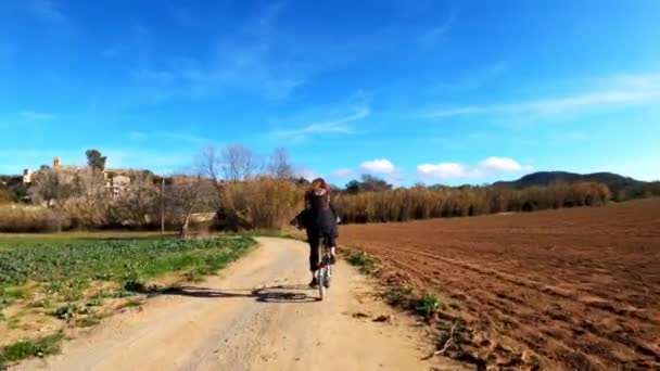 Una niña de pelo largo monta en bicicleta en un camino rural entre campos, bosques y prados. Un paisaje pintoresco. Paseo en bicicleta rural. Callejuelas, bosques con pinos y cipreses. Luz dorada . — Vídeo de stock