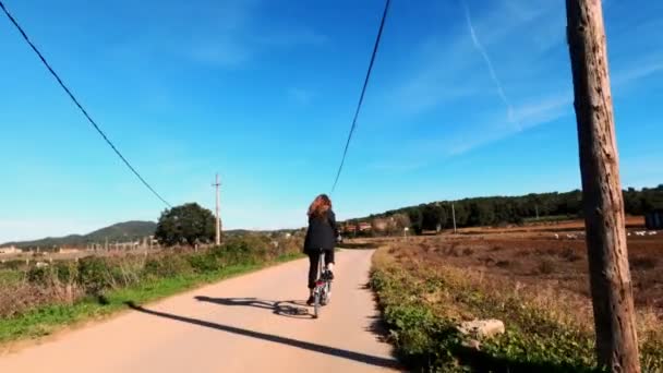 Una ragazza con i capelli lunghi va in bicicletta su una strada rurale tra campi, boschi e prati. Pittoresca campagna. Bicicletta rurale. Alleys, boschi di pini e cipressi. Luce dorata . — Video Stock