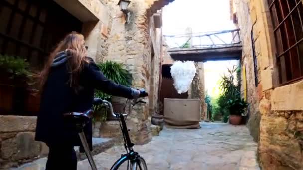 Una chica con el pelo largo pasea con una bicicleta en el pequeño pueblo medieval de piedra. Un paisaje pintoresco. Paseo en bicicleta rural. Calle de piedra vieja. Luz dorada . — Vídeos de Stock