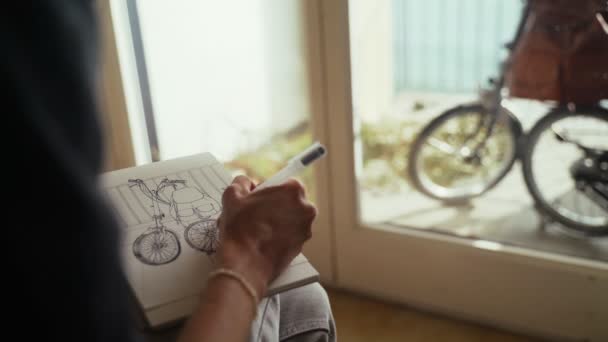 De kunstenaar maakt schetsen. Meisje tekent een fiets in een schetsboek. Zwart-wit tekening door voering op wit papier. Close-up om te schetsen. Gouden zonlicht. Handtekening. Tekenen en tekenen. — Stockvideo