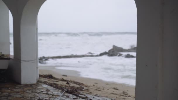 Utsikt över det stormiga havet genom de vita valven i en liten stad. Tittar på stormen. En liten by under en storm. Svårt rasande hav. Stora vågor. Vindstarkt regnigt väder. Utsikt över det dramatiska havet. — Stockvideo