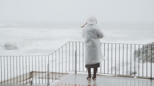 A girl in a white raincoat is walking along the stormy sea. A woman is watching the storm. Young woman walking on the seaside. Severe stormy sea. Big waves. Strong wind. Windy rainy weather. — 비디오
