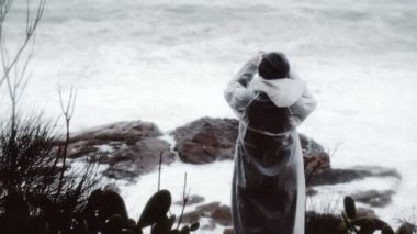 Beyaz yağmurluklu bir kız fırtınalı denizde yürüyor. Bir kadın fırtınayı izliyor. Deniz kenarında yürüyen genç bir kadın. Şiddetli fırtınalı deniz. Büyük dalgalar. Güçlü rüzgar. Rüzgarlı yağmurlu hava.