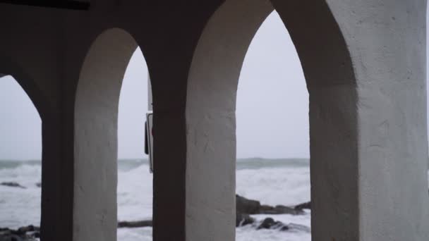 Vista do mar tempestuoso através dos arcos brancos de uma pequena cidade. A ver a tempestade. Pequena aldeia durante uma tempestade. Mar em fúria severa. Ondas grandes. Tempo ventoso e chuvoso. Vista do oceano dramático . — Vídeo de Stock