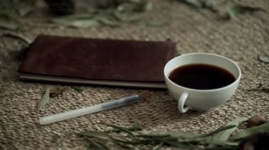 Hasır hasırlı kompozisyon, kuru bitkiler, bir defter ve bir fincan kahve. Bir fincan kahve tutan kadın ellerinin yakın çekimi. Kahve içiyorum. Doğal renkler. Kahvaltıda. Çevre dostu estetik..