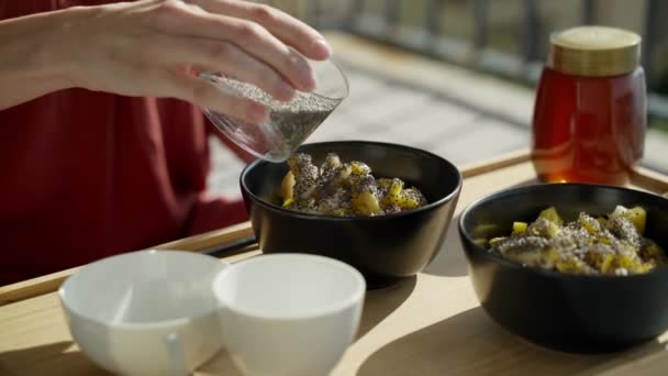 Colazione sulla terrazza. Picnic sulla terrazza soleggiata. Colazione sana. La ragazza aggiunge semi di chia alla farina d'avena. Saporito porridge sano con noci, frutta e miele. Tè mattutino. Prodotti ecologici. Colazione dietetica — Video Stock