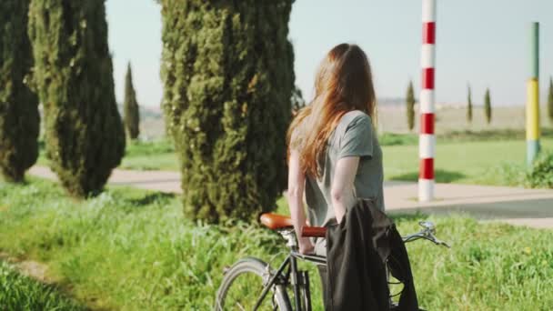 Mujer joven descansando apoyada en una bicicleta. Una chica mira un hermoso paisaje de primavera. Descansa en un callejón de cipreses. Deportes al aire libre. Estilo de vida saludable. Paisaje rural. Colores de primavera. Luz dorada . — Vídeos de Stock