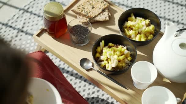 Frukost på terrassen. Picknick på solterrassen. En hälsosam frukost. Flicka tillför frukt till havregryn. Smaklig hälsosam gröt med nötter, frukt och honung. Morgonte. Ekologiska produkter. Kostfrukost. — Stockvideo