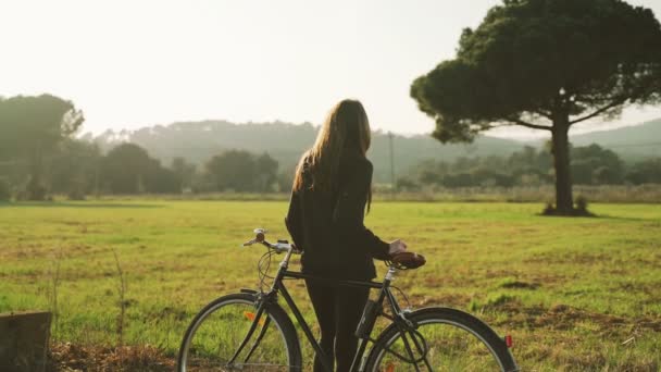 Mujer joven descansando apoyada en una bicicleta. Una chica mira un hermoso paisaje de primavera. Árbol solitario y hermosas colinas. Deportes al aire libre. Estilo de vida saludable. Paisaje rural. Colores de primavera. Luz dorada . — Vídeo de stock