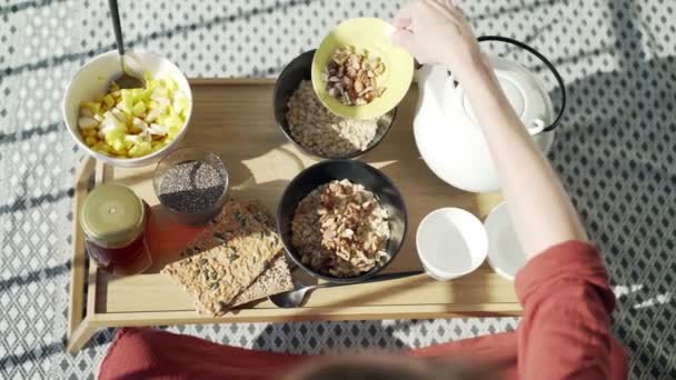 Frühstück auf der Terrasse. Picknick auf der sonnigen Terrasse. Gesundes Frühstück. Mädchen fügt Haferflocken Nüsse hinzu. Leckerer gesunder Brei mit Nüssen, Früchten und Honig. Morgentee. Öko-Produkte. Diätfrühstück. — Stockvideo