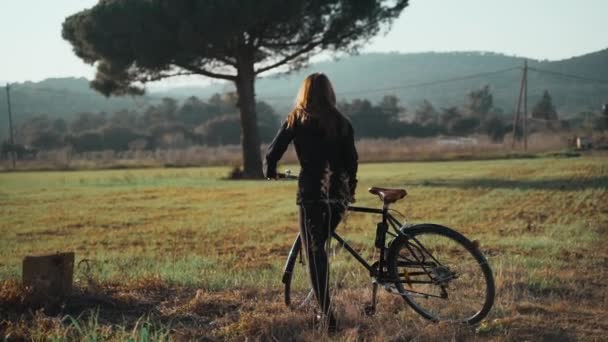 Μια νεαρή γυναίκα περπατάει με ένα ποδήλατο κατά μήκος ενός αγροτικού δρόμου. Όμορφο ανοιξιάτικο τοπίο. Μοναχικό δέντρο και όμορφοι λόφοι. Υπαίθρια αθλήματα. Υγιεινός τρόπος ζωής Αγροτικό τοπίο. Ανοιξιάτικα χρώματα Χρυσό φως. — Αρχείο Βίντεο