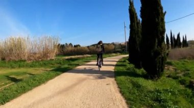 Uzun saçlı bir kız kırsal bir yolda bisiklet sürüyor tarlalar, ormanlar ve çayırlar arasında. Resimli kırsal kesim. Kırsal bisiklet yolculuğu. Sırt çantalı kız. Sokaklar, ormanlar ve selvi ağaçları. Altın ışık.