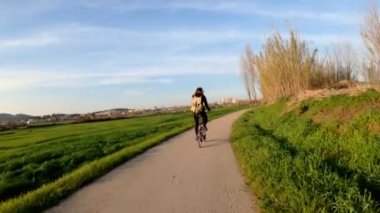 Uzun saçlı bir kız kırsal bir yolda bisiklet sürüyor tarlalar, ormanlar ve çayırlar arasında. Resimli kırsal kesim. Kırsal bisiklet yolculuğu. Sırt çantalı kız. Sokaklar, ormanlar ve selvi ağaçları. Altın ışık.