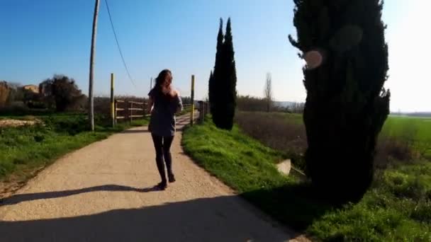 Jonge atletische vrouw aan het joggen op een lenteochtend. Joggen langs de weg tussen cipressen en groene velden. Buitensporten. Een gezonde levensstijl. Prachtig landelijk landschap. Voorjaarskleuren. Lopen. — Stockvideo