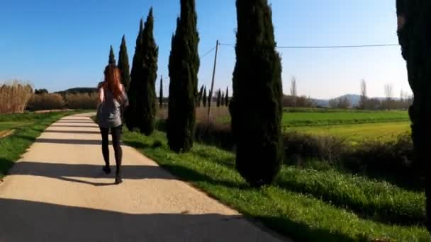 Mujer atlética joven corriendo en una mañana de primavera. Correr a lo largo de la carretera entre cipreses y campos verdes. Deportes al aire libre. Estilo de vida saludable. Hermoso paisaje rural. Colores de primavera. Correr. . — Vídeos de Stock