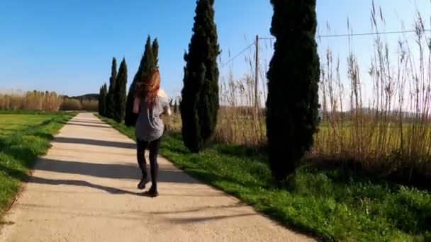 Ung atletisk kvinna joggar en vårmorgon. Joggar längs vägen bland cypresser och gröna fält. Utomhussporter. En hälsosam livsstil. Vacker landsbygd. Vårfärger. Löpning. — Stockvideo