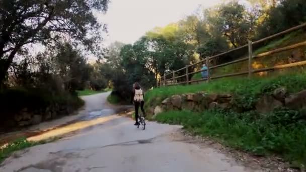 Una ragazza con i capelli lunghi va in bicicletta su una strada rurale tra campi, boschi e prati. Pittoresca campagna. Bicicletta rurale. Ragazza con lo zaino. Alleanze, foreste e cipressi. Luce dorata . — Video Stock