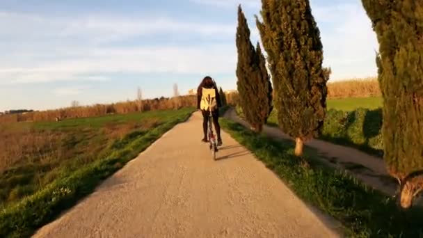 Una ragazza con i capelli lunghi va in bicicletta su una strada rurale tra campi, boschi e prati. Pittoresca campagna. Bicicletta rurale. Ragazza con lo zaino. Alleanze, foreste e cipressi. Luce dorata . — Video Stock