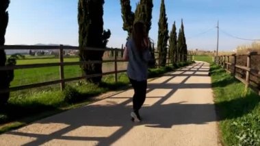 Bir bahar sabahı koşan genç bir atletik kadın. Yol boyunca, selvi ağaçları ve yeşil tarlalar arasında koşuyor. Açık hava sporları. Sağlıklı bir yaşam tarzı. Güzel kırsal manzara. Bahar renkleri. Çalışıyor.