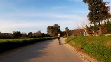 Uzun saçlı bir kız kırsal bir yolda bisiklet sürüyor tarlalar, ormanlar ve çayırlar arasında. Resimli kırsal kesim. Kırsal bisiklet yolculuğu. Sırt çantalı kız. Sokaklar, ormanlar ve selvi ağaçları. Altın ışık.