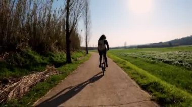 Uzun saçlı bir kız kırsal bir yolda bisiklet sürüyor tarlalar, ormanlar ve çayırlar arasında. Resimli kırsal kesim. Kırsal bisiklet yolculuğu. Sırt çantalı kız. Sokaklar, ormanlar ve selvi ağaçları. Altın ışık.