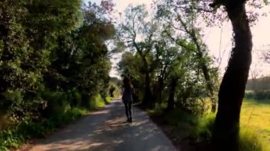 Bir bahar sabahı koşan genç bir atletik kadın. Yol boyunca ağaçlar ve yeşil tarlalar arasında koşuyor. Açık hava sporları. Sağlıklı bir yaşam tarzı. Güzel kırsal manzara. Bahar renkleri. Altın ışık. Çalışıyor.