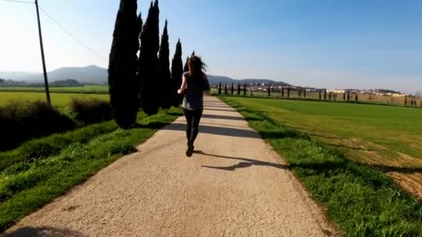 Jeune femme athlétique faisant du jogging un matin de printemps. Jogging le long de la route entre les cyprès et les champs verts. Sports de plein air. Mode de vie sain. Beau paysage rural. Couleurs printanières. Courir . — Video