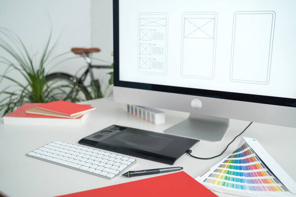 Web designer at work. Graphic designer. Mobile application design. Designer desktop. UX, UI design. Thumbnails of a mobile application on a computer screen. Gadgets, sketchbook and phone on the table
