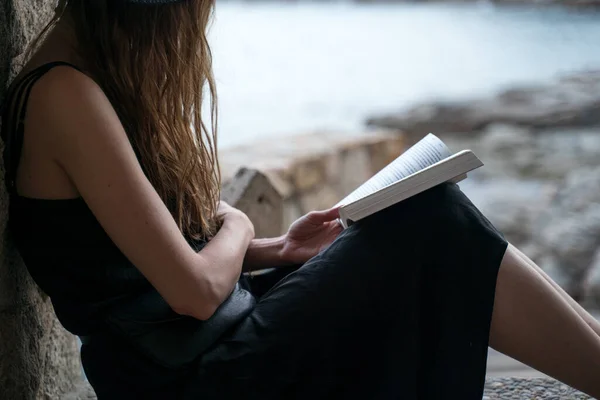 Meisje leest boek. Een meisje met een zwarte hoed en een zwarte zijden jurk zit in de nis van een vissershut aan de kust en leest een boek. Zeezicht op een achtergrond. Buiten studeren, een roman lezen. — Stockfoto