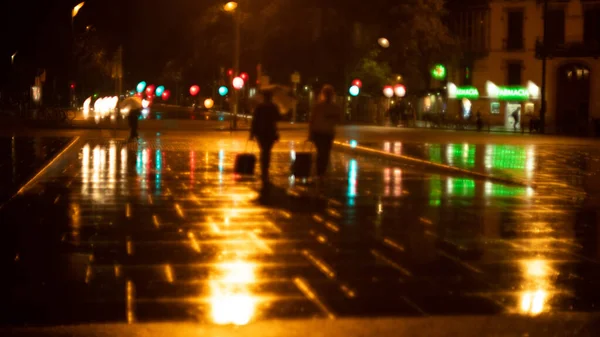 Rainy evening in the city. Blurry figures of people walking. Shiny raindrops on the street. Multi-colored city lights in the background. Creative evening lights and blurred shadows. Twilight mood.