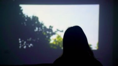 Duvardaki ekranın arka planında bir kızın silueti. Kadın projektörle film izliyor. Evde ne yapacağım? Evde film izliyordum. Evde kal. Evde iyi eğlenceler. Karantina. 