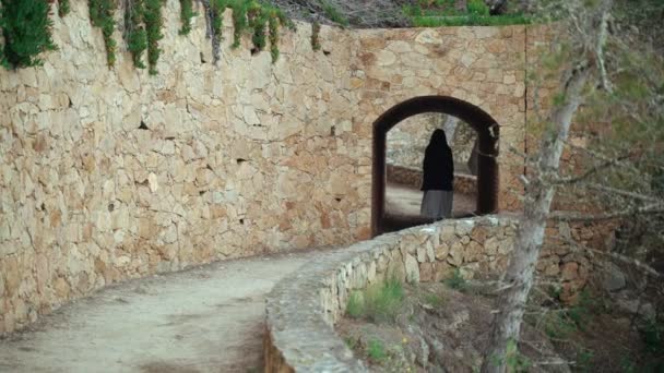 Chica con un vestido de rayas blancas y chaqueta gris camina a lo largo del camino costero y pasa a través de un túnel de piedra. Mujer de pelo largo pasea por el hermoso campo. Luz dorada en un paisaje idílico . — Vídeos de Stock